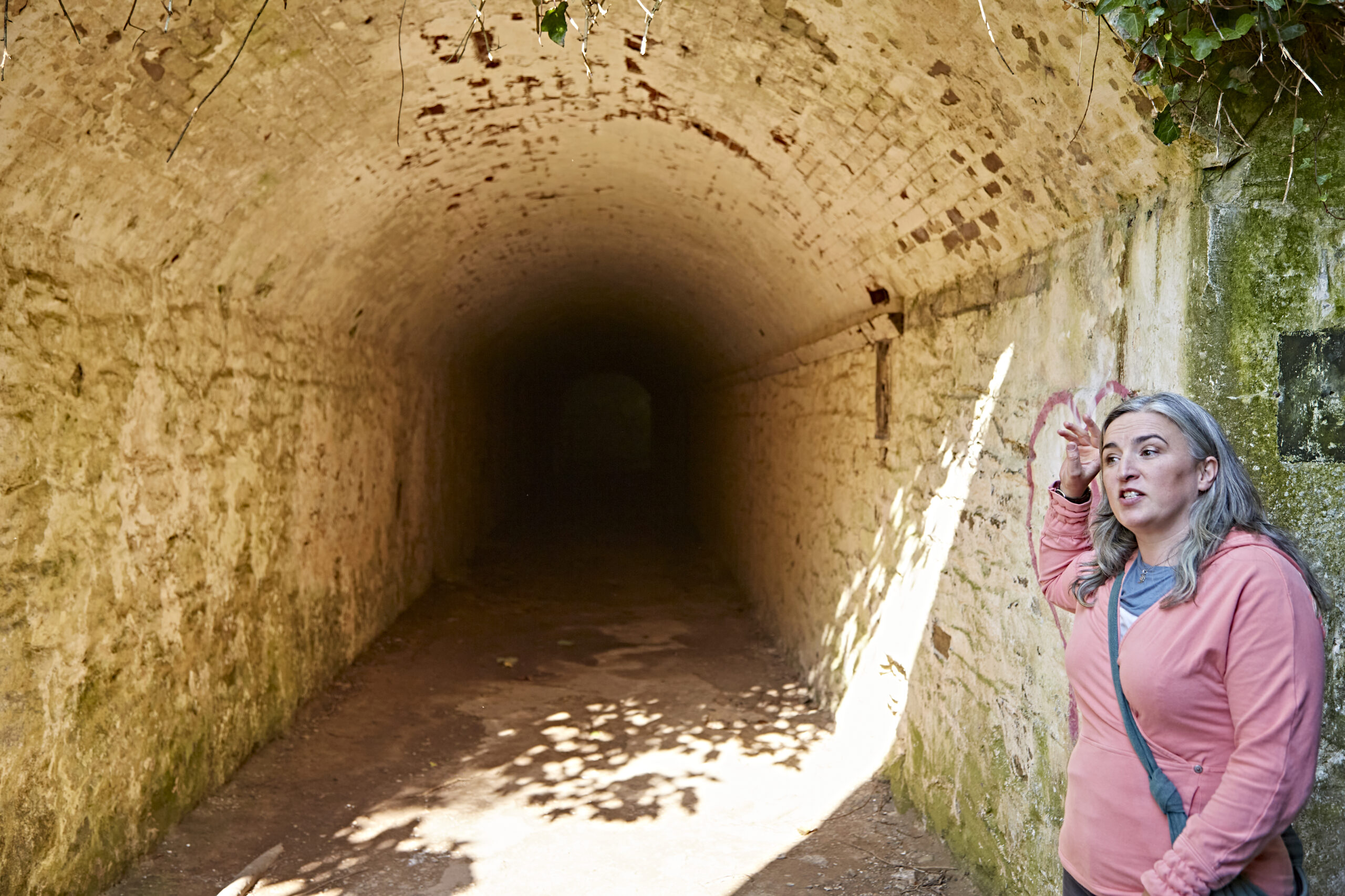 A photo of the owner of Feather and Stone standing at the end of a stone tunnel. Nothing can be seen in the tunnel past the first five metres. Anne is talking and has her right hand raised to the height of her head as if she is about to point.